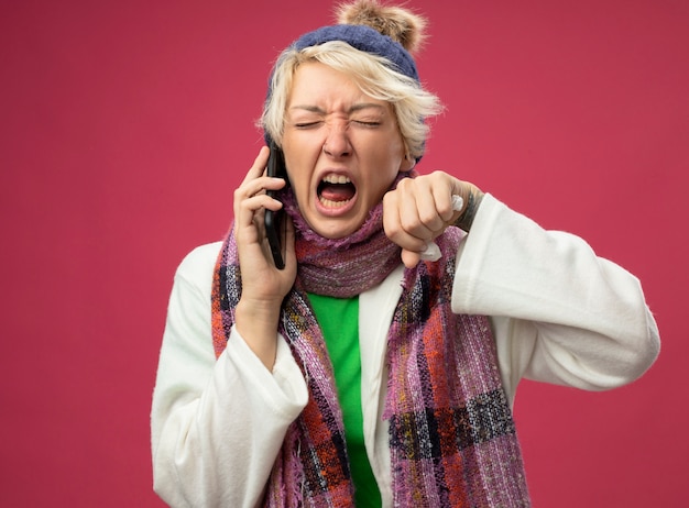 Donna malsana malata frustrata arrabbiata con capelli corti in sciarpa calda e cappello sensazione di malessere gridando mentre parla al telefono cellulare in piedi sopra il muro rosa