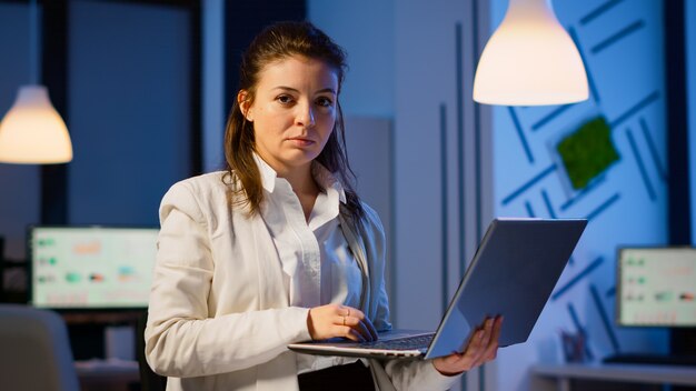 Donna manager con laptop, digitando e guardando soddisfatto la telecamera in piedi in una start-up a tarda notte. Dipendente concentrato che utilizza la rete tecnologica wireless facendo gli straordinari per la lettura del lavoro.