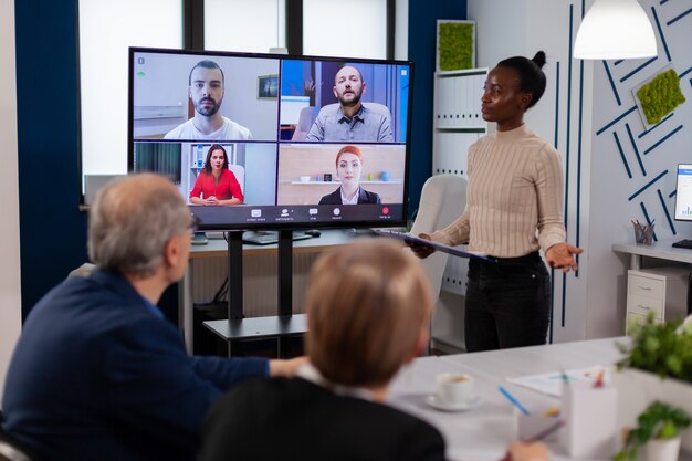 Donna manager nera che parla con colleghi in remoto in videochiamata sullo schermo della tv, presentando nuovi partner commerciali