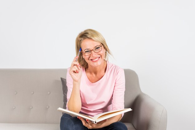 Donna matura bionda sorridente che si siede sulla penna e sul libro di tenuta del sofà contro il contesto bianco