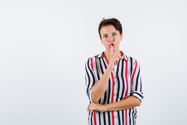 Donna matura in camicia a righe che mostra gesto di silenzio e che sembra seria, vista frontale.