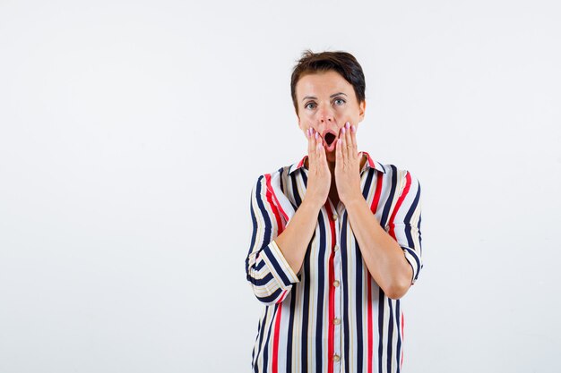 Donna matura in camicia a righe tenendo le mani vicino alla bocca, tenendo la bocca aperta e guardando sorpreso, vista frontale.