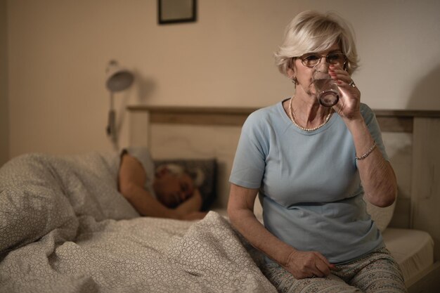 Donna matura seduta sul letto e bevendo un bicchiere d'acqua mentre suo marito dorme sullo sfondo