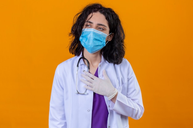 donna medico che indossa camice bianco con lo stetoscopio nella mascherina protettiva medica alla ricerca di malessere in piedi con la mano sul petto su arancione isolato