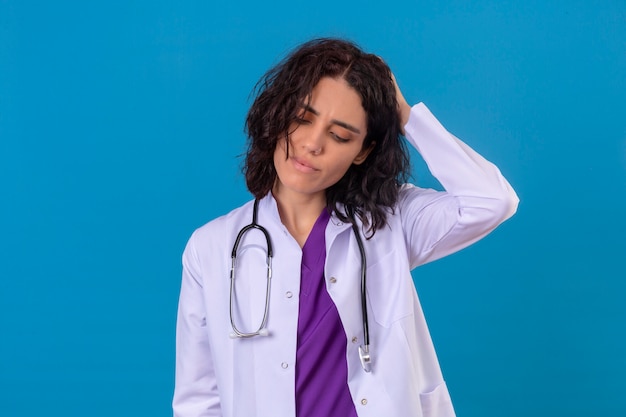 donna medico che indossa camice bianco con stetoscopio cercando incerto con dubbio pensando con la mano sulla testa in piedi sul blu isolato