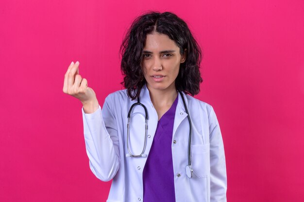 donna medico che indossa camice bianco con stetoscopio facendo gesto di denaro con le mani per chiedere il pagamento dello stipendio in piedi sul rosa isolato