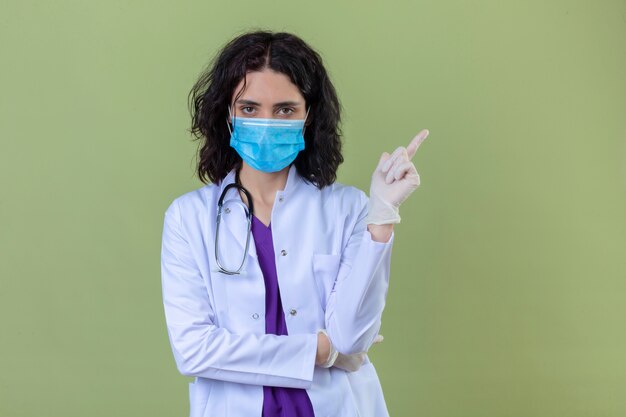 donna medico che indossa camice bianco con stetoscopio in maschera protettiva medica con viso serio che punta con il dito a lato in piedi sul verde isolato