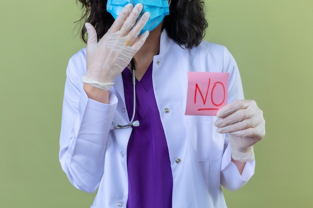 donna medico che indossa camice bianco con stetoscopio in maschera protettiva medica tenendo carta promemoria senza parola che sembra sorpreso sul verde isolato