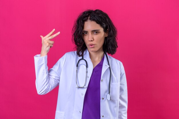 donna medico che indossa camice bianco con stetoscopio sorridente fiducioso mostrando il numero due con le dita in alto sul rosa isolato