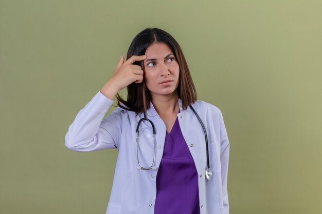 donna medico che indossa camice bianco e con lo stetoscopio in piedi con il dito contro il tempio pensando cercando di fare una scelta sul verde isolato