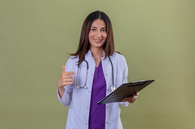 donna medico che indossa camice bianco e con stetoscopio azienda appunti sorridente fiducioso che punta ad esso con il dito indice in piedi sul verde isolato