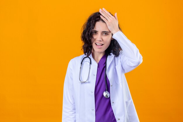 Donna medico che indossa mantello bianco con uno stetoscopio in piedi con la mano sulla testa per errore ricorda l'errore dimenticato cattivo concetto di memoria in piedi su arancione isolato