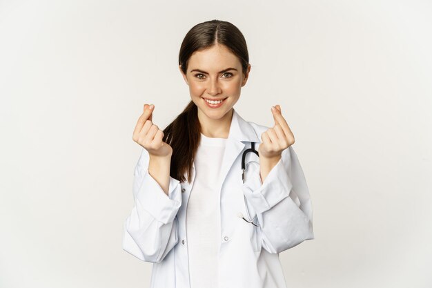 Donna medico che mostra i cuori delle dita sorridenti con cura in piedi in uniforme su sfondo bianco