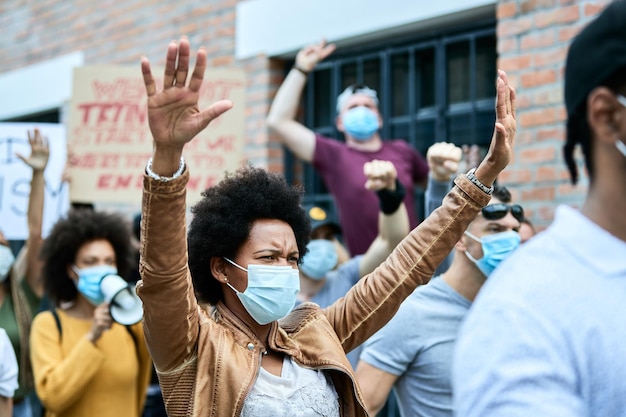 Donna nera dispiaciuta che indossa una maschera protettiva mentre protesta con la folla di persone per le strade della città durante l'epidemia di COVID19