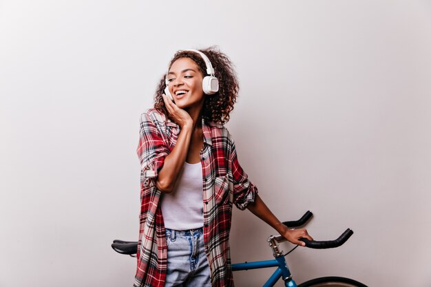 Donna nera gioiosa ascoltando musica e posa con la bicicletta. Tiro al coperto di accattivante signora con i capelli ondulati camicia sorridente su bianco