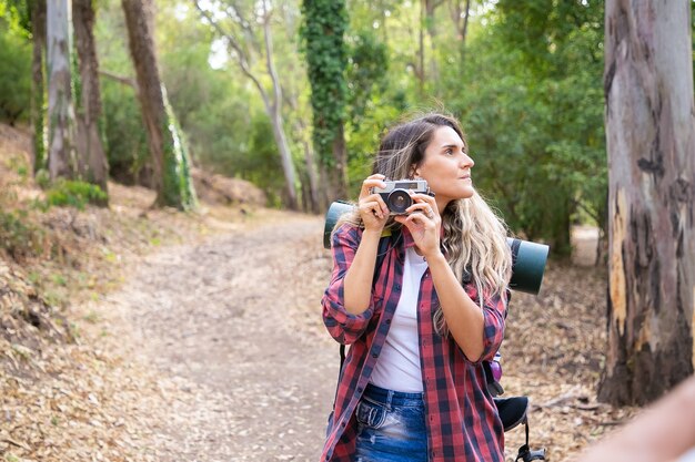 Donna pensierosa che tiene la macchina fotografica, guardandosi intorno e in piedi sulla strada. Turista femminile esplorando la natura e prendendo foto della foresta. Concetto di turismo, avventura e vacanze estive