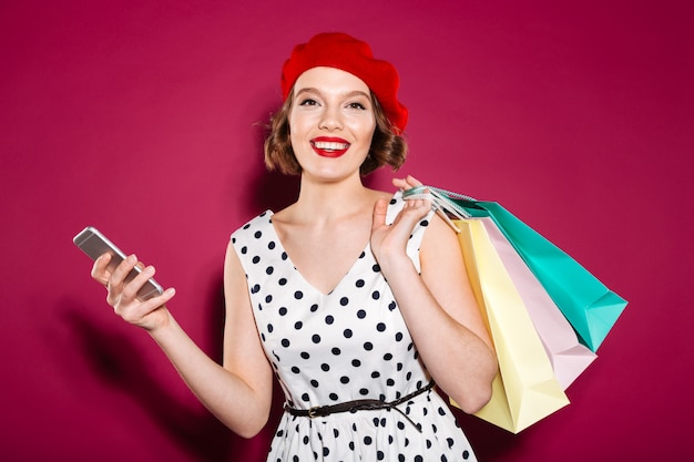 Donna piacevole dello zenzero in pacchetti e smartphone della tenuta del vestito mentre esaminando la macchina fotografica sopra il rosa