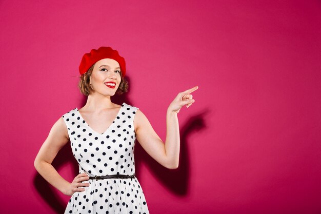 Donna piacevole dello zenzero in vestito con il braccio sull'anca che indica il copyspace e che esamina la macchina fotografica sopra il rosa