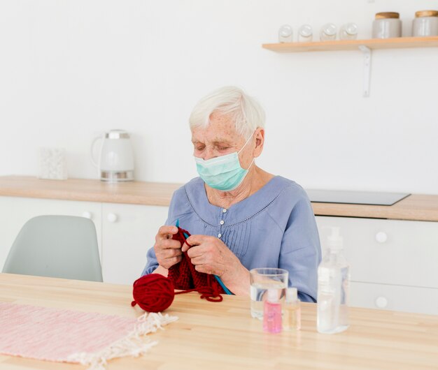 Donna più anziana con la maschera medica che tricotta a casa