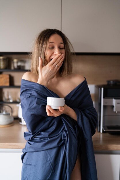 Donna positiva assonnata avvolta in un lenzuolo blu che beve caffè al mattino in cucina