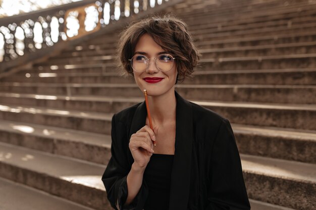 Donna positiva con capelli corti e labbra luminose in vetri che sorridono all'esterno. Signora alla moda in abiti neri in posa sulle scale.