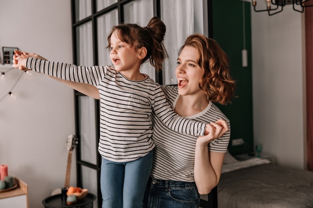 Donna positiva e sua figlia in magliette a righe ridono e ballano in appartamento.