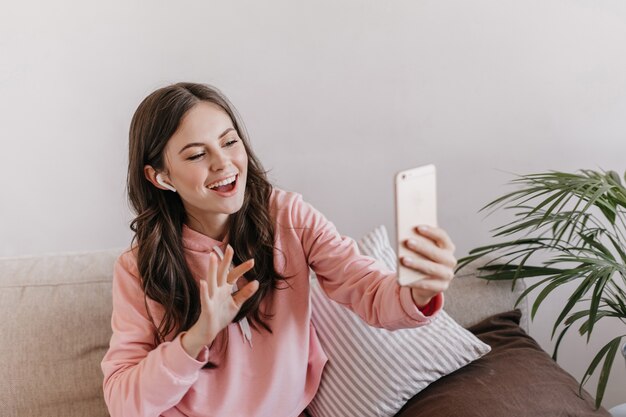 Donna positiva in abito sportivo rosa parlando al telefono in cuffie wireless e seduto sul divano