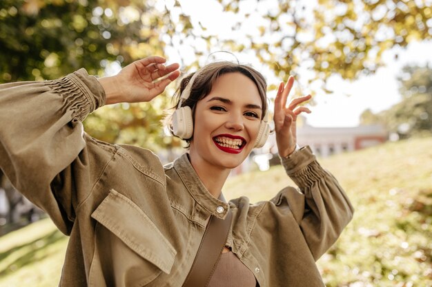 Donna positiva in giacca di jeans verde oliva e cuffie bianche che sorridono all'esterno. La donna dai capelli corti con le labbra rosse si diverte all'aria aperta.