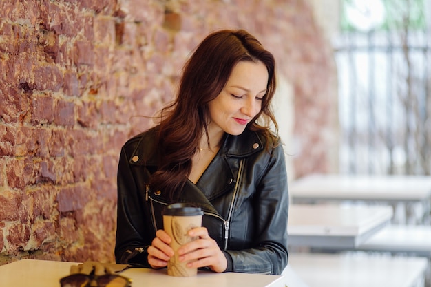 Donna positiva in vestiti alla moda che bevono bevande da asporto mentre è seduto al tavolo sulla strada vicino al caffè durante il giorno