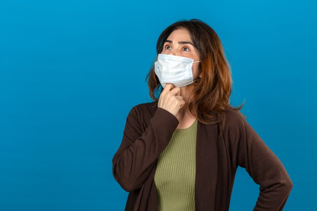 Donna premurosa che indossa cardigan marrone nella maschera protettiva medica che sta con la mano sul mento che cerca pensante sopra la parete blu isolata