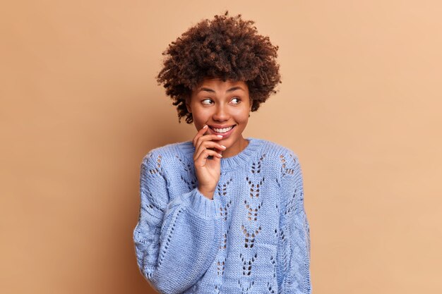 Donna premurosa sognante con capelli afro distoglie lo sguardo sorrisi dolcemente concentrato da parte sorride vestito positivamente in pose di ponticello blu contro il muro marrone