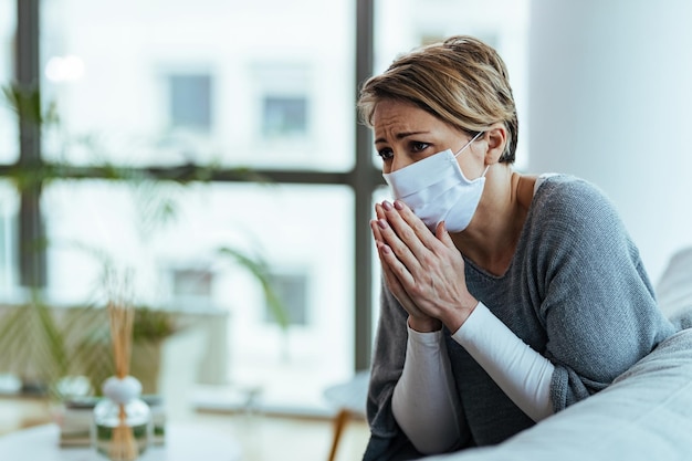 Donna preoccupata che prega con le mani giunte mentre indossa una maschera protettiva e si sente triste durante la pandemia di coronavirus