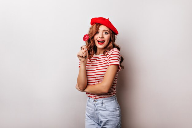 Donna riccia allegra che mangia lecca-lecca sul muro bianco. Affascinante ragazza francese in berretto in posa con la caramella.