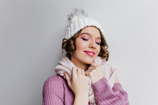 donna riccia bianca felice in maglione di lana. Ritratto dell'interno di bella ragazza con trucco rosa che posa in cappello e sciarpa in una giornata fredda.