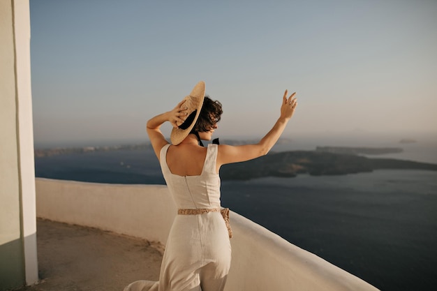 Donna riccia bruna in elegante abito beige e cappello a tesa larga di paglia gode di un clima soleggiato e di una splendida vista sul mare