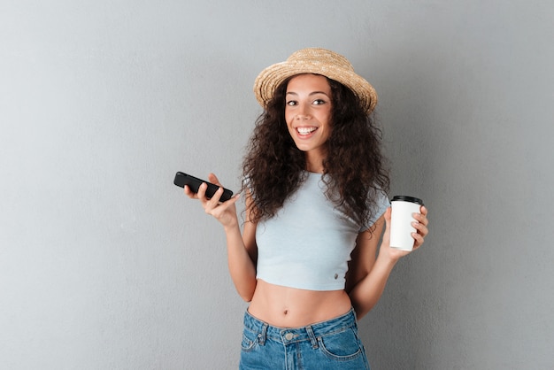 Donna riccia felice in cappello con smartphone e caffè