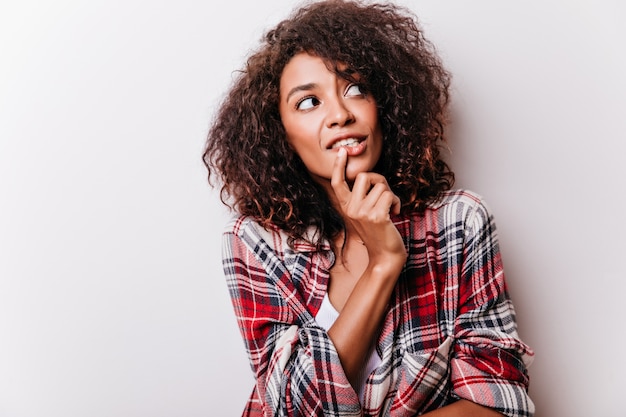 Donna riccia pensierosa guardandosi intorno e toccandosi il mento. modello femminile africano in camicia a scacchi isolata sulla luce.