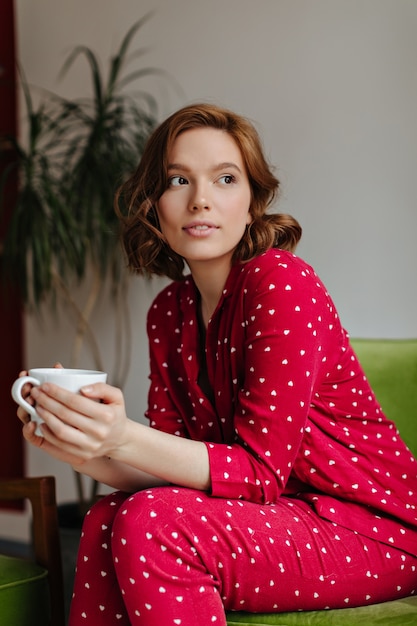 Donna riccia sognante che tiene tazza di caffè e distoglie lo sguardo. Tiro al coperto di allegra giovane signora in pigiama rosso che beve il tè.