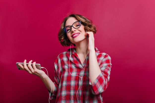 Donna romantica con un sorriso soddisfatto ascoltando musica in auricolari. Ritratto dell'interno del modello femminile bianco allegro in vetri con l'acconciatura corta in piedi sul muro bordeaux.