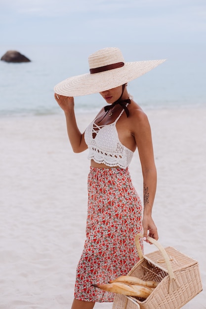 Donna romantica sulla spiaggia in gonna lavorata a maglia e cappello di paglia che tiene il cestino con pane Eco life