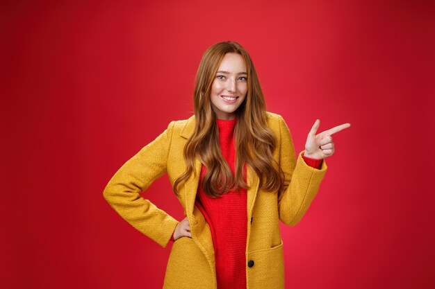 Donna rossa attraente elegante e sicura di sé con le lentiggini in cappotto esterno giallo che punta a destra con la pistola del dito e sorride gioiosamente come macchina fotografica mentre mostra una fantastica promozione sul muro rosso.