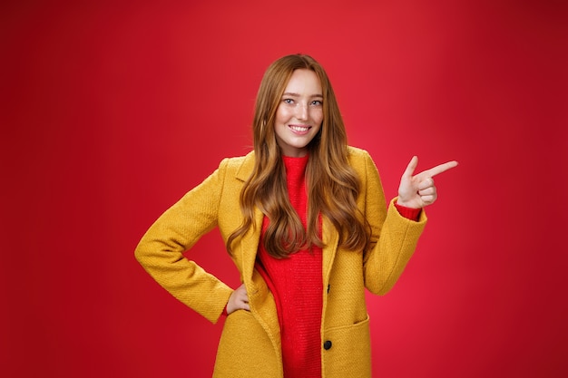 Donna rossa attraente elegante e sicura di sé con le lentiggini in cappotto esterno giallo che punta a destra con la pistola del dito e sorride gioiosamente come macchina fotografica mentre mostra una fantastica promozione sul muro rosso.