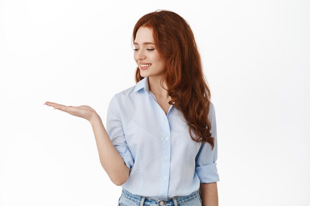 Donna rossa carina che guarda il palmo aperto e sorride al prodotto, tenendo in mano lo spazio della copia dell'oggetto, in piedi in camicetta da ufficio su sfondo bianco