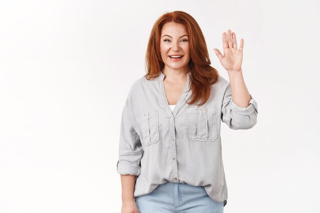 Donna rossa di mezza età che viene incontro dicendo ciao a tutti conoscere squadra alzare la mano salutando amichevole ciao gesto sorridente a trentadue denti accogliente figlia amici in piedi ottimista muro bianco