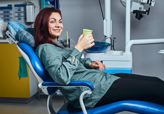 Donna rossa felice che si siede su una sedia del dentista che tiene una tazza con il colluttorio in una clinica.