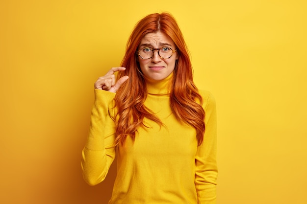 donna rossa infelice fa un piccolo gesto dimostra qualcosa di minuscolo vestito con un maglione casual.