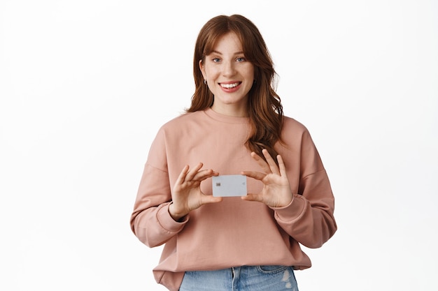 donna rossa sorridente che mostra la carta di credito, consiglia la banca, in piedi in abiti casual su bianco