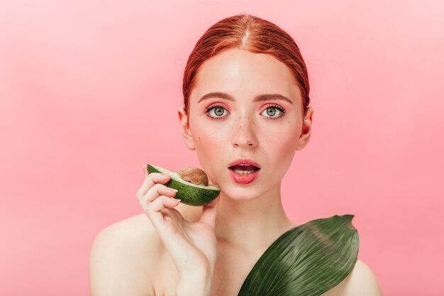 Donna scioccata che tiene avocado e che guarda l'obbiettivo. Ragazza graziosa dello zenzero con cibo sano che esprime stupore su sfondo rosa.