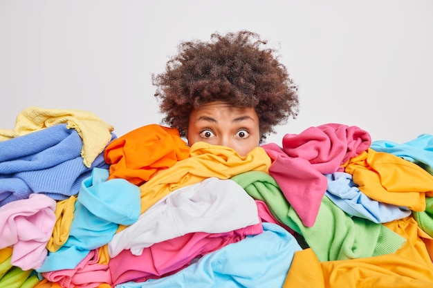 Donna scioccata con i capelli ricci afro fissa gli occhi spiaccicati annegati in un'enorme pila di vestiti colorati pulisce l'armadio seleziona i vestiti per la donazione o il riciclaggio bianco