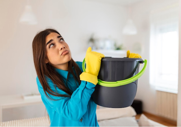 Donna scioccata guarda il soffitto mentre raccoglie l'acqua che perde nel soggiorno di casa Donna preoccupata che tiene il secchio mentre le goccioline d'acqua perdono dal soffitto nel soggiorno